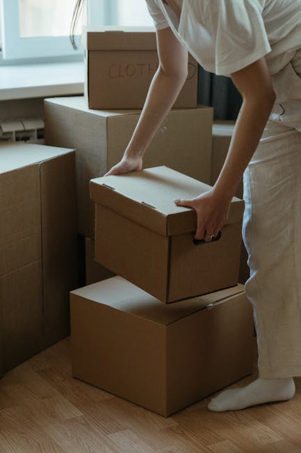 A person wearing light-colored clothing is inside a property, stacking medium-sized cardboard boxes that are sealed with packing tape. The boxes are arranged on the floor near a window, with some labeled, including one marked 'CLOTHES' on top of a larger box. The individual is preparing the boxes for home relocation, carefully placing one box onto another, demonstrating the packing and lifting process involved in furniture transport. In the background, more packed boxes are visible, along with a neutral-toned interior. The scene is well-lit with natural light coming through the window, and a wooden floor is visible underneath. This image exemplifies the packing phase of house removals, a key service offered by Removals London, supporting efficient and organized moving logistics for clients in the London area.