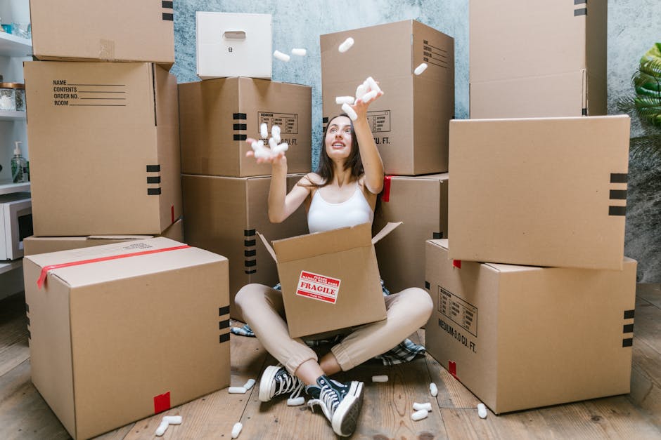 A young woman sitting cross-legged on a wooden floor inside a room during a home relocation process is surrounded by numerous cardboard moving boxes of various sizes, some sealed with red packing tape and others with black printed labels. She is smiling and appears to be playfully tossing white packing peanuts into the air, with some falling into an open box resting on her lap. Behind her, larger boxes are stacked against a blue textured wall, and a small part of a white kitchen appliance and a potted plant are visible to the left and right, respectively. The setting is well-lit, with natural or artificial lighting illuminating the scene, highlighting the materials used in packing, such as cardboard and plastic wrap, and the actions involved in furniture transport or packing during the process of house removals. This image exemplifies the movement and packing activities typical of home relocation services offered by Removals London, as referenced on the N1 moving tips for Islington studio removals page.