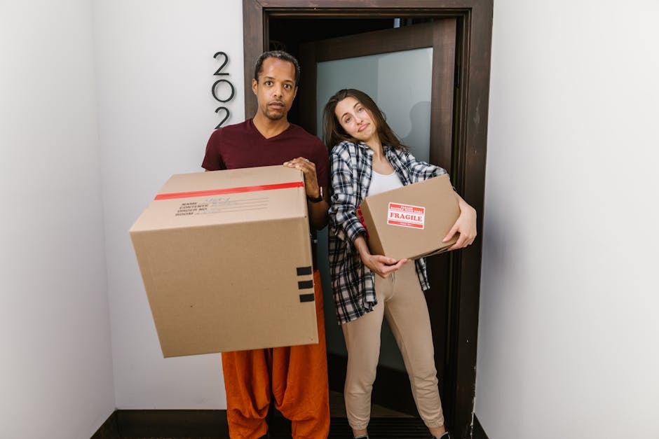 A person wearing light-colored clothing is inside a property, stacking medium-sized cardboard boxes that are sealed with packing tape. The boxes are arranged on the floor near a window, with some labeled, including one marked 'CLOTHES' on top of a larger box. The individual is preparing the boxes for home relocation, carefully placing one box onto another, demonstrating the packing and lifting process involved in furniture transport. In the background, more packed boxes are visible, along with a neutral-toned interior. The scene is well-lit with natural light coming through the window, and a wooden floor is visible underneath. This image exemplifies the packing phase of house removals, a key service offered by Removals London, supporting efficient and organized moving logistics for clients in the London area.