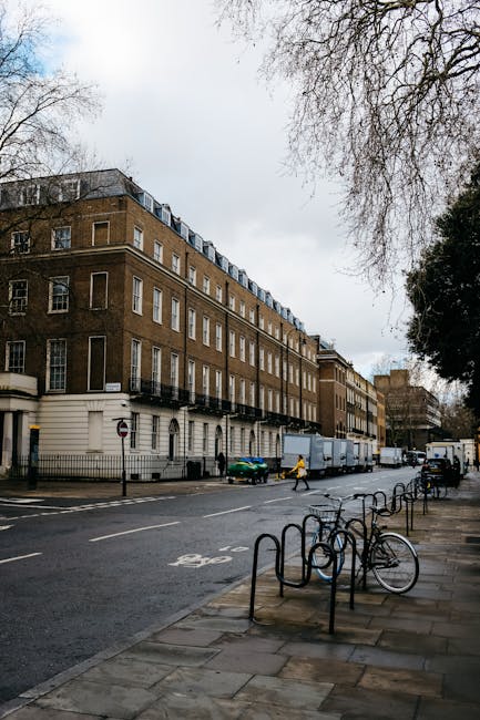 A daytime street scene outside a multi-storey residential building with a brown brick façade and white window frames, situated in Chelsea’s SW3 area. The building features several rows of windows and small balconies along the upper floors. On the pavement in front, a row of black metal bicycle racks is visible, with a couple of bicycles parked. Several large cardboard boxes and wrapped furniture pieces are staged outside the building, indicating a home relocation process. Moving blankets and plastic wrapping are used to protect furniture during transport. A small green car and a larger white moving truck are parked parallel to the curb, with the truck positioned beside the entrance for loading purposes. Two individuals are actively engaged in loading or unloading items from the truck, reflecting a furniture transport and packing and moving operation. The scene is illuminated by natural daylight, with overhanging branches of trees partially visible in the upper right corner, and a pedestrian crossing can be seen further along the street, supporting the urban relocation environment managed by Removals London.