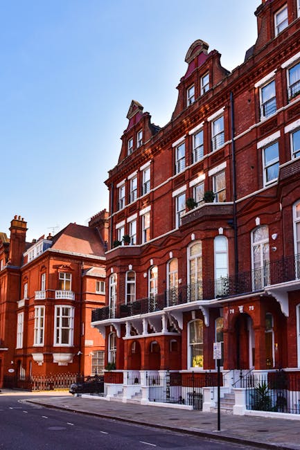 The image depicts a row of red-brick Victorian-style residential buildings in Chelsea, London, with several large windows, some with white frames and decorative brickwork. The building in the foreground features a white-painted ground floor with an entrance, a small staircase, and a wrought-iron balcony on the first floor. Inside a ground-floor apartment, a furniture trolley loaded with cardboard boxes, plastic-wrapped household items, and furniture is visible near the doorway, indicating a home relocation process. Several cardboard boxes of different sizes are stacked on the pavement outside, some secured with packing tape and wrapped in clear plastic. A moving blanket and lifting straps are used to assist with the loading process. The scene is set under clear daylight, with a blue sky above, and the street appears quiet, consistent with a residential area. Removals London, a professional moving company, is engaged in loading furniture and packed boxes into a white moving van parked adjacent to the property, in preparation for transportation to a new location as part of a comprehensive furniture transport and packing service aligned with house removals in Chelsea.