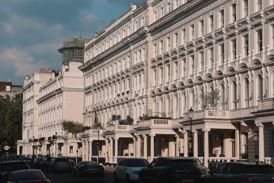 A daytime street scene outside a multi-storey residential building with a brown brick façade and white window frames, situated in Chelsea’s SW3 area. The building features several rows of windows and small balconies along the upper floors. On the pavement in front, a row of black metal bicycle racks is visible, with a couple of bicycles parked. Several large cardboard boxes and wrapped furniture pieces are staged outside the building, indicating a home relocation process. Moving blankets and plastic wrapping are used to protect furniture during transport. A small green car and a larger white moving truck are parked parallel to the curb, with the truck positioned beside the entrance for loading purposes. Two individuals are actively engaged in loading or unloading items from the truck, reflecting a furniture transport and packing and moving operation. The scene is illuminated by natural daylight, with overhanging branches of trees partially visible in the upper right corner, and a pedestrian crossing can be seen further along the street, supporting the urban relocation environment managed by Removals London.